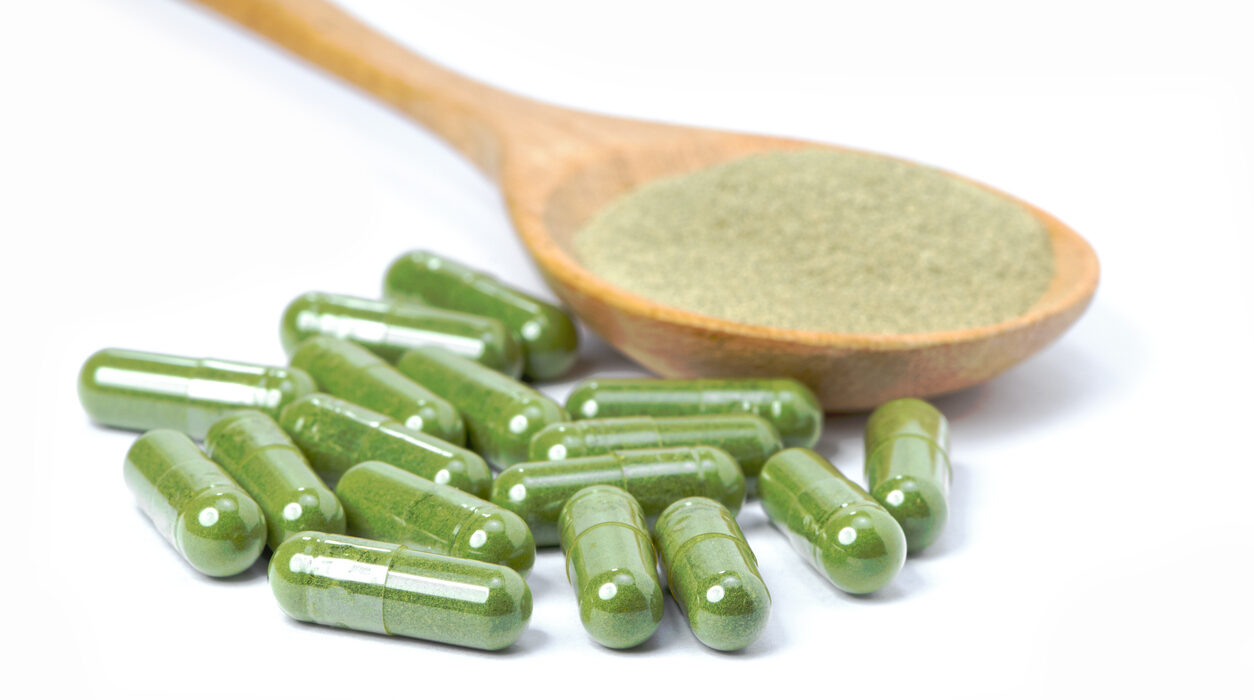 Green herbal powder capsule and dried leaf plant powder in wooden spoon isolated on white background.